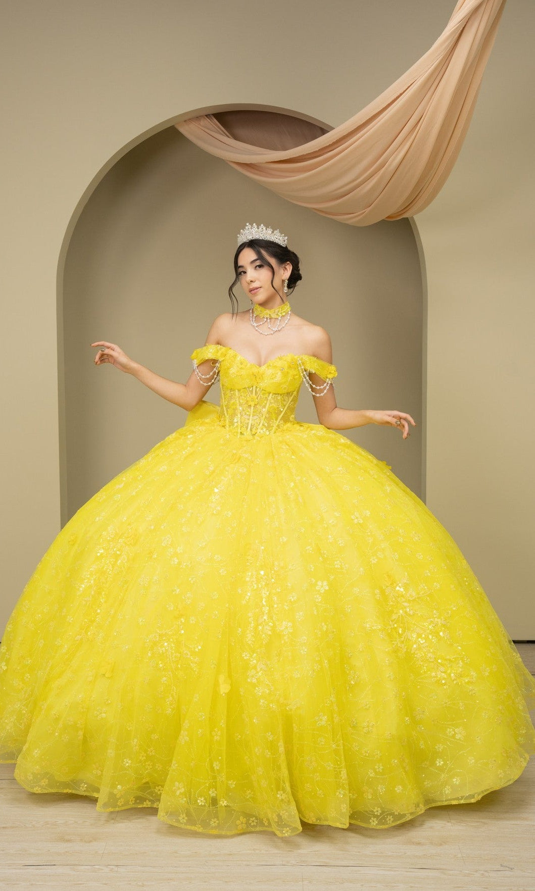 A woman in the yellow Quinceanera Dress 8415JQ by Cinderella Couture and a tiara stands before an arched, fabric-draped backdrop, embodying elegance.